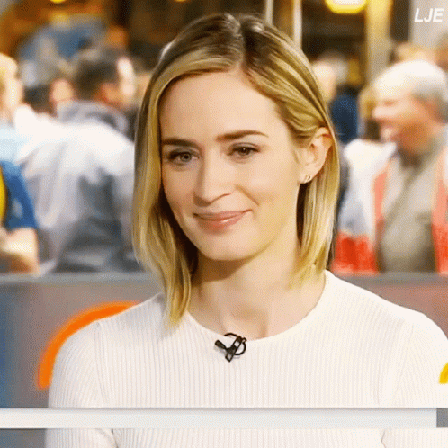 A Woman Wearing A White Sweater With A Microphone Around Her Neck Looks At The Camera GIF