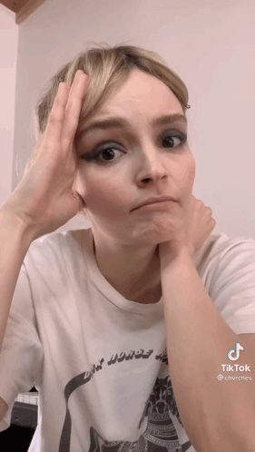 A Woman Wearing A White T-Shirt Has Her Hand On Her Forehead . GIF