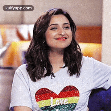 A Woman Wearing A White T-Shirt With A Heart On It Is Smiling And Sitting On A Couch . GIF