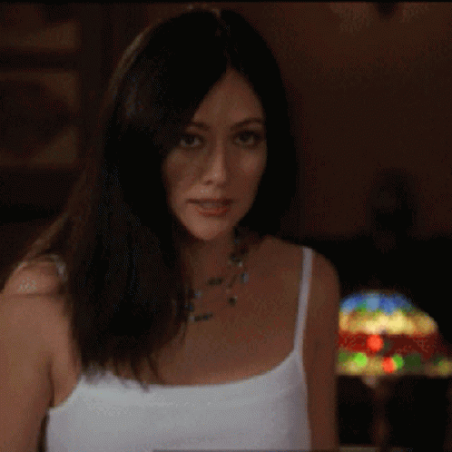 A Woman Wearing A White Tank Top And A Necklace Is Looking At The Camera GIF