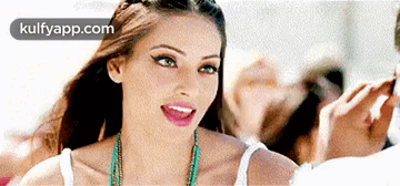 A Woman Wearing A White Tank Top And A Turquoise Necklace Smiles For The Camera . GIF