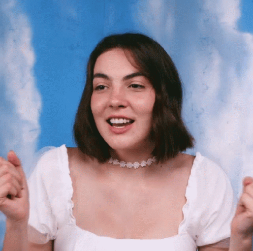 A Woman Wearing A White Top And A Choker Smiles With Her Hands In The Air GIF