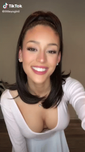 A Woman Wearing A White Top With A Plunging Neckline Is Smiling . GIF