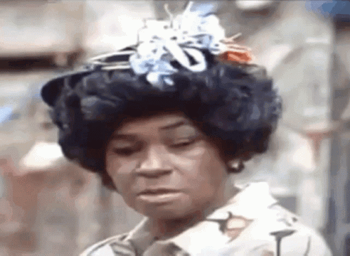 A Woman Wearing A Wig And A Hat With Flowers On It Is Making A Funny Face . GIF