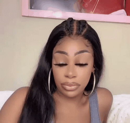 A Woman Wearing A Wig And Hoop Earrings Is Sitting On A Bed . GIF