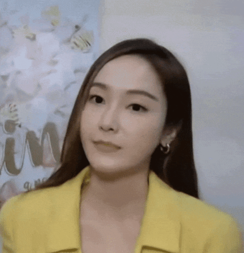 A Woman Wearing A Yellow Jacket And Earrings Is Looking At The Camera GIF