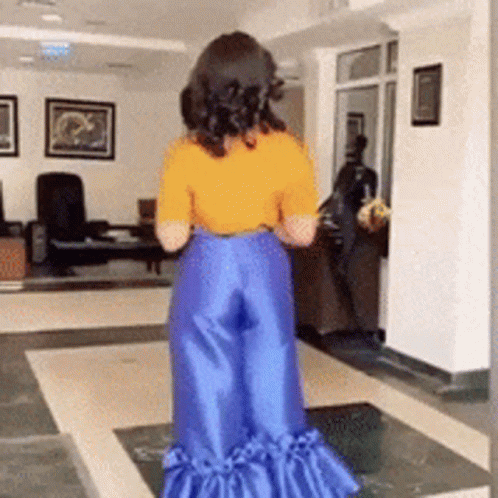A Woman Wearing A Yellow Shirt And Blue Pants Is Standing In A Living Room GIF