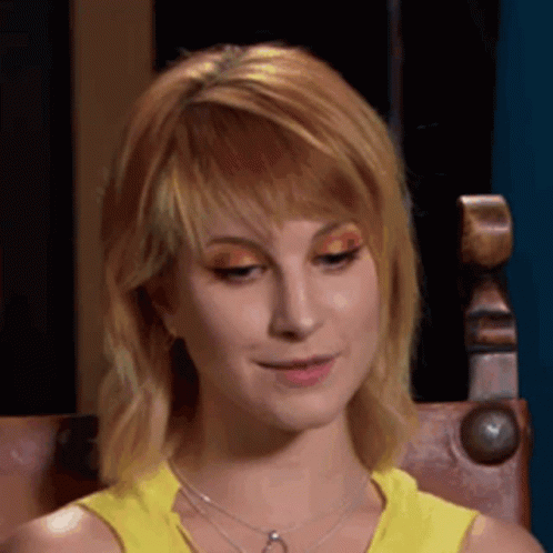 A Woman Wearing A Yellow Tank Top And A Necklace Is Sitting In A Chair And Smiling . GIF