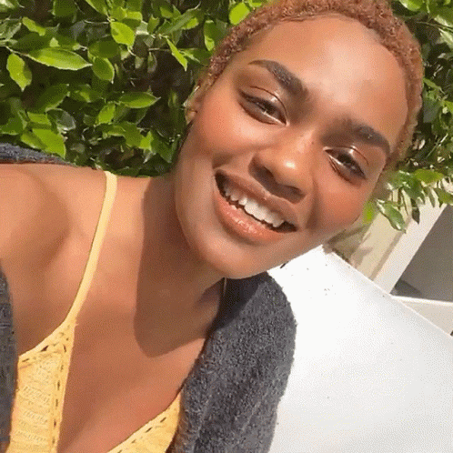 A Woman Wearing A Yellow Top And A Gray Sweater Smiles For The Camera GIF