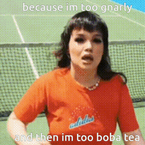 A Woman Wearing An Orange Adidas Shirt Is Standing In Front Of A Tennis Court GIF