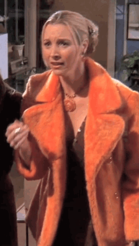 A Woman Wearing An Orange Fur Coat Is Standing In A Room With Other People . GIF
