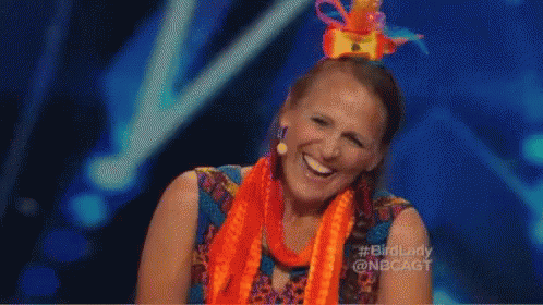 A Woman Wearing An Orange Scarf And A Colorful Hat Is Laughing On A Stage . GIF