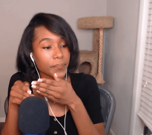 A Woman Wearing Ear Buds Is Talking Into A Microphone In Front Of A Cat Tree GIF