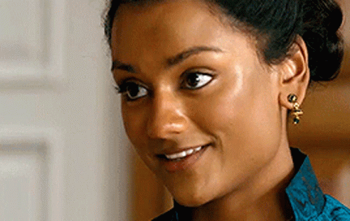 A Woman Wearing Earrings And A Blue Shirt Smiles GIF