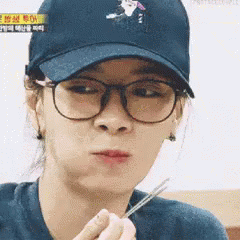 A Woman Wearing Glasses And A Baseball Cap Is Holding Chopsticks In Her Hand . GIF