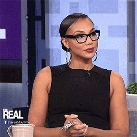 A Woman Wearing Glasses And A Black Tank Top Is Sitting In Front Of A Sign That Says Real GIF