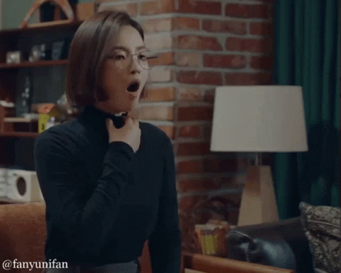 A Woman Wearing Glasses And A Black Turtleneck Is Yawning In A Living Room GIF