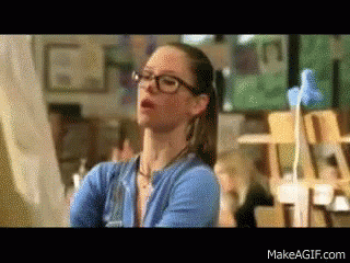 A Woman Wearing Glasses And A Blue Sweater Is Standing In Front Of A Painting GIF