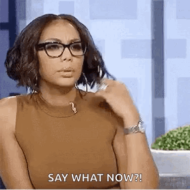 A Woman Wearing Glasses And A Brown Tank Top Is Sitting Down And Saying `` Say What Now '' . GIF