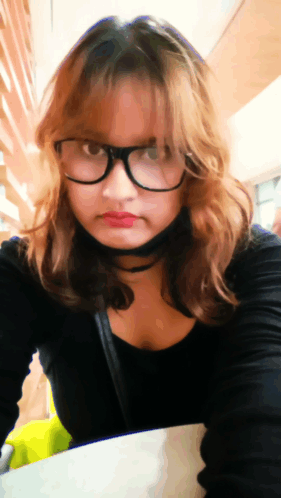 A Woman Wearing Glasses And A Choker Is Sitting At A Table GIF