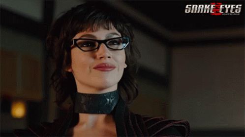 A Woman Wearing Glasses And A Choker With A Snake Eyes Logo Behind Her GIF
