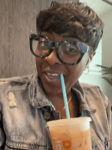 A Woman Wearing Glasses And A Denim Jacket Drinks Through A Straw From A Cup That Has The Letter D On It GIF