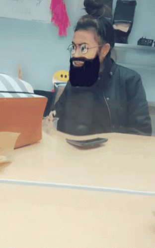 A Woman Wearing Glasses And A Fake Beard Sits At A Table GIF