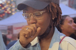 A Woman Wearing Glasses And A Hat Coughs GIF