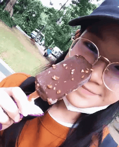 A Woman Wearing Glasses And A Hat Eating A Chocolate Bar GIF