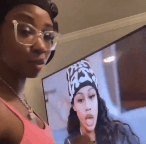 A Woman Wearing Glasses And A Hat Is Standing In Front Of A Television Screen . GIF