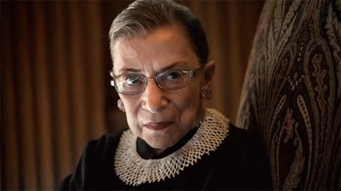 A Woman Wearing Glasses And A Knitted Collar Looks At The Camera GIF