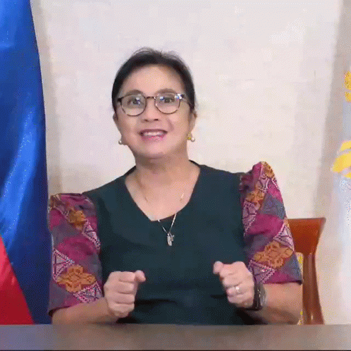A Woman Wearing Glasses And A Necklace Is Sitting At A Desk With Her Hands In The Air . GIF