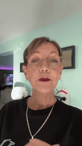 A Woman Wearing Glasses And A Necklace Looks At The Camera While Making A Face GIF