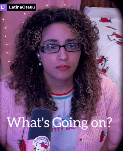 A Woman Wearing Glasses And A Pink Hello Kitty Sweater Says What 'S Going On In Front Of A Microphone GIF