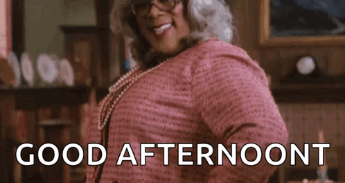 A Woman Wearing Glasses And A Pink Shirt Is Standing In A Living Room And Smiling . GIF