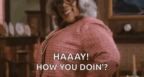 A Woman Wearing Glasses And A Pink Shirt Is Standing In A Living Room And Smiling . GIF