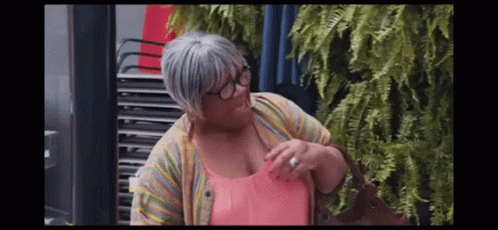 A Woman Wearing Glasses And A Pink Shirt Is Standing In Front Of A Stack Of Chairs . GIF