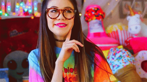 A Woman Wearing Glasses And A Tie Dye Shirt Is Standing In Front Of A Pile Of Stuffed Animals . GIF