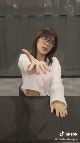 A Woman Wearing Glasses And A White Shirt Is Sitting At A Table With A Sign That Says Wee GIF