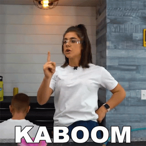 A Woman Wearing Glasses And A White Shirt Stands In Front Of A Sign That Says " Kaboom " GIF