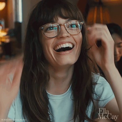 A Woman Wearing Glasses And A White Shirt With Ella Mccay On The Bottom Right GIF