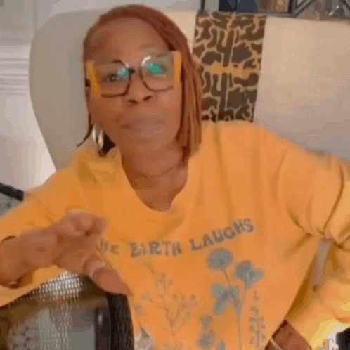 A Woman Wearing Glasses And A Yellow Sweater Is Sitting In A Chair And Making A Funny Face . GIF