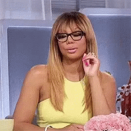 A Woman Wearing Glasses And A Yellow Top Is Sitting In A Chair . GIF