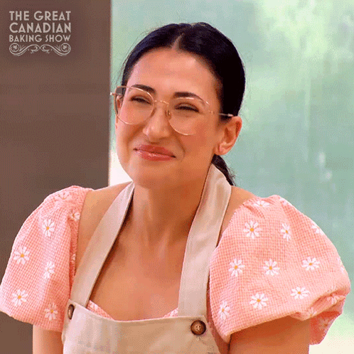 A Woman Wearing Glasses And An Apron Is Smiling In Front Of The Great Canadian Baking Show Logo GIF