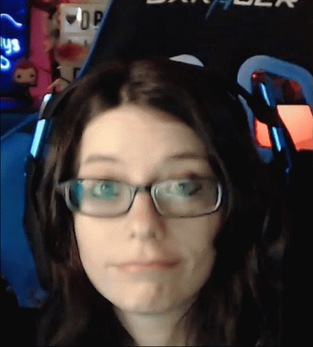 A Woman Wearing Glasses And Headphones Is Making A Face In Front Of A Sign That Says Dxracer GIF