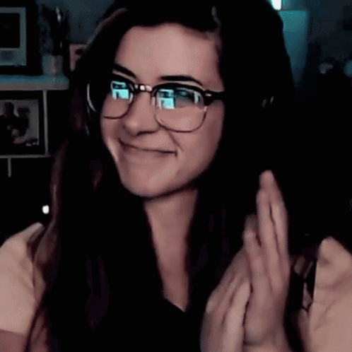 A Woman Wearing Glasses And Headphones Is Smiling And Waving GIF