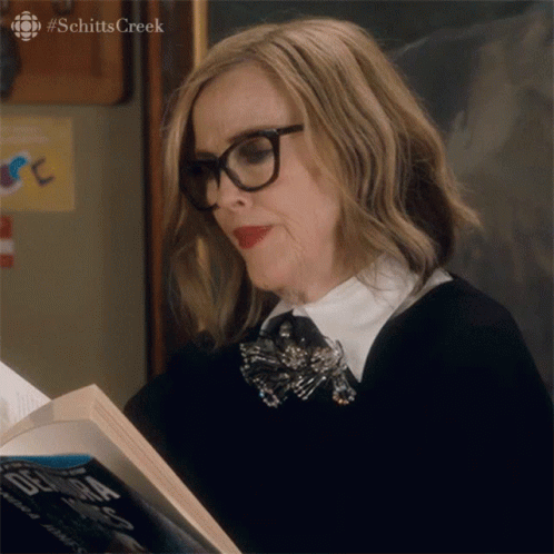 A Woman Wearing Glasses Is Reading A Book From Schitts Creek . GIF