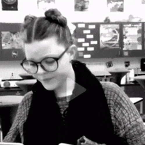 A Woman Wearing Glasses Is Sitting At A Desk In A Classroom . GIF
