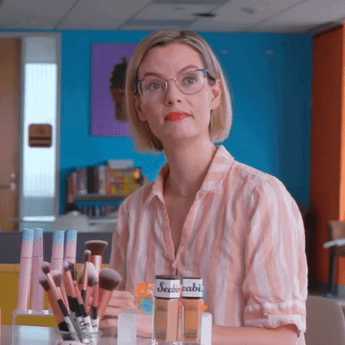 A Woman Wearing Glasses Is Sitting At A Table With Makeup Brushes And A Bottle Of Sealedabi GIF