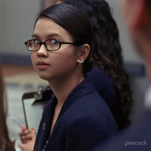 A Woman Wearing Glasses Is Sitting In Front Of A Clipboard With Peacock Written On The Bottom GIF